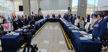 Salón de convenciones con personas de pie, mesas con manteles azules, cámara, micrófonos y un cartel que dice 'Comisión de Población'
