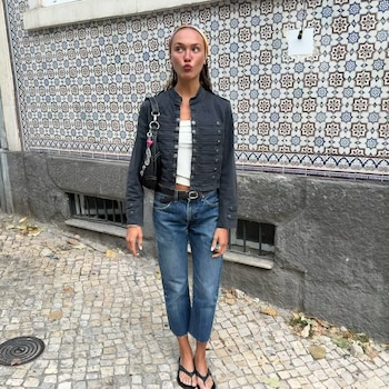 Mujer joven con chaqueta oscura, camiseta blanca, pantalón vaquero, sandalias de dedo y diadema. Muro de azulejos y calle empedrada