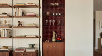 Vista de una estantería de madera con libros y objetos decorativos junto a un nicho con azulejos rojos, estantes de cristal, vasos y un jarrón.