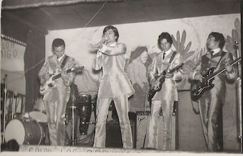 Fotografía en blanco y negro de cuatro músicos en un escenario; uno canta al micrófono, tres tocan guitarras eléctricas, vestidos con trajes brillantes