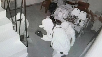 Tres personas con el rostro cubierto caminan dentro de una habitación junto a una escalera y varios muebles