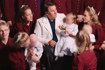 Un hombre de mediana edad con chaqueta blanca y camisa azul está sentado, rodeado de varias mujeres y niños pequeños vestidos con túnicas y tiaras