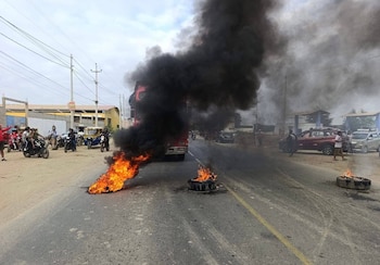 Una carretera bloqueada con tres neumáticos en llamas que emiten humo negro. Un camión y motocicletas están detenidos, con personas a los lados