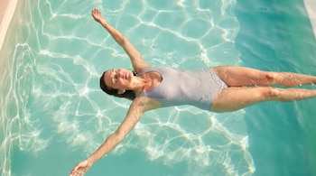 Persona con traje de baño gris flotando de espaldas y con los brazos abiertos en una piscina iluminada por el sol.