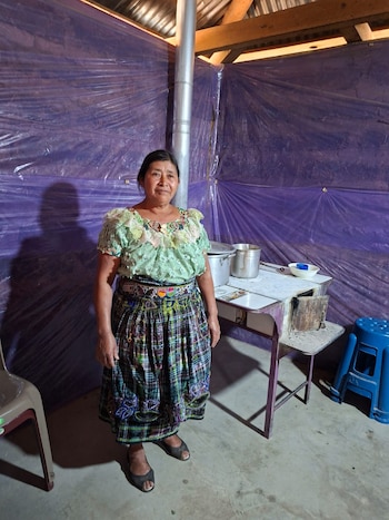 Una mujer de pie con blusa verde bordada y falda a cuadros. Un fogón de metal con ollas, pared de plástico morado, techo de chapa y piso de cemento