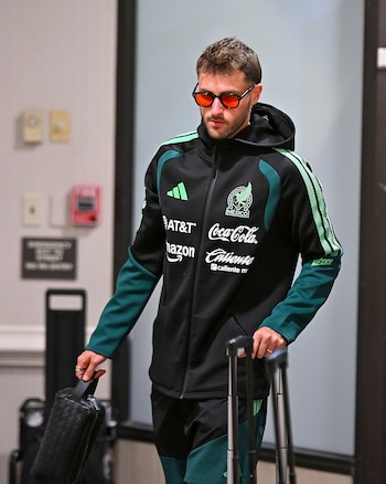 Un futbolista con chaqueta deportiva negra de la Selección de México, gafas de sol y maletas camina por un pasillo interior