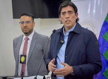 Dos hombres, uno de ellos hablando ante un micrófono con camisa azul y chaqueta oscura, y otro con traje gris y corbata roja, en una sala de conferencias