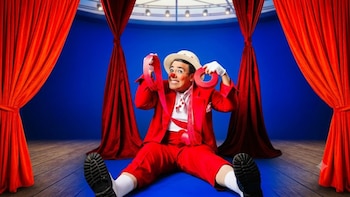 Hombre disfrazado de payaso con traje rojo, nariz roja y sombrero beige, sentado en un suelo azul entre cortinas rojas, sujetando cinta roja