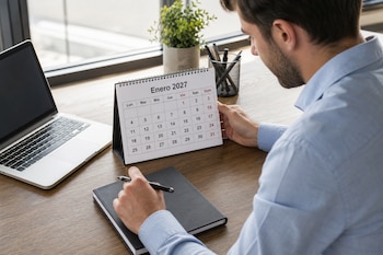 Hombre en un escritorio de oficina consultando un calendario de enero de 2027, junto a un ordenador portátil y un cuaderno.