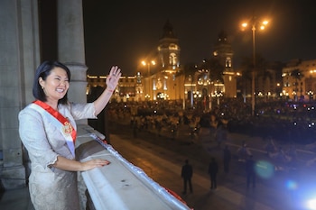 Mujer de pelo corto y sonrisa, vestida de beige con banda roja y blanca, saluda desde balcón de piedra. Multitud y edificios iluminados de noche al fondo