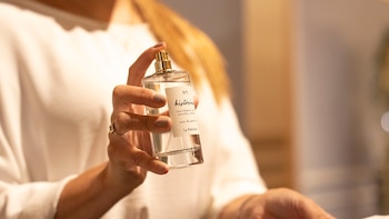 Primer plano de manos sosteniendo una botella de perfume transparente con pulverizador dorado y etiqueta que dice 'Nº 5 historias' y detalles