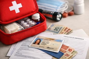 Un botiquín rojo abierto con vendajes y pastillas, junto a una identificación, billetes de dinero y un formulario de contactos de emergencia.