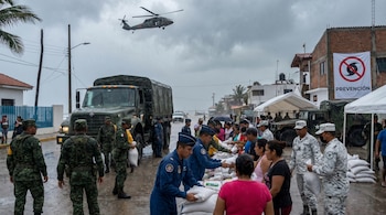 Helicóptero militar sobrevuela mientras elementos del Ejército, Fuerza Aérea y Guardia Nacional de México distribuyen sacos de arena a civiles bajo la lluvia en una zona costera.