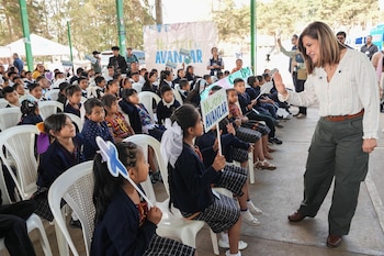 Una mujer de cabello castaño y camisa blanca se acerca a un grupo de niños sentados en sillas blancas, algunos con uniformes, sosteniendo pequeños carteles al aire libre