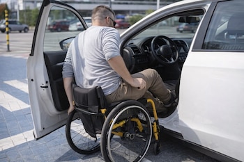 Hombre con gafas de sol sentado en una silla de ruedas amarilla y negra al lado de un coche blanco con la puerta abierta.