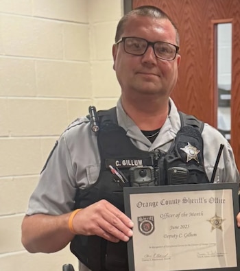 Hombre uniformado, C. Gillum, con gafas, sonríe a la cámara mientras sostiene un certificado de "Oficial del Mes" de la Oficina del Sheriff del Condado de Orange