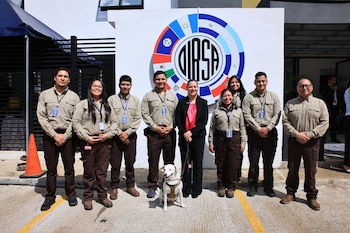 Ocho personas, siete en uniforme y una de traje, posan al aire libre con un perro blanco delante de una pared blanca con un gran logo circular de DIASA