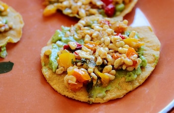 Primer plano de una tostada con base de tortilla de maíz, cubierta con guacamole cremoso, granos blancos y trozos de pimiento rojo y amarillo