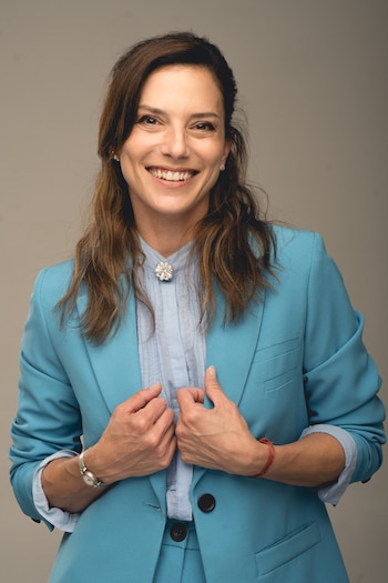 Manuela Pal sonríe a cámara con una chaqueta azul turquesa y camisa celeste. Lleva un broche de flor, un reloj plateado y una pulsera roja