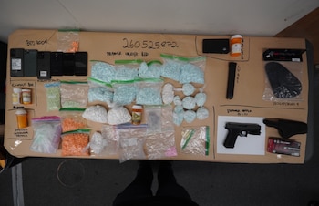 Múltiples bolsas con pastillas de colores, frascos de farmacia, teléfonos, una pistola negra y munición sobre una mesa con texto manuscrito