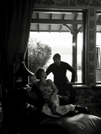 Fotografía en blanco y negro de una mujer sentada en un sofá y un hombre en el alféizar de una ventana. Interior con cortinas y papel tapiz