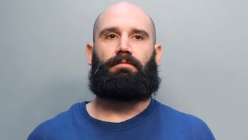Mugshot de un hombre calvo con barba oscura frondosa y camiseta azul, mirando fijamente a la cámara con un fondo gris claro