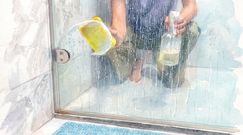 Una persona agachada sostiene una esponja amarilla y un pulverizador frente a un cristal de ducha con gotas de agua.