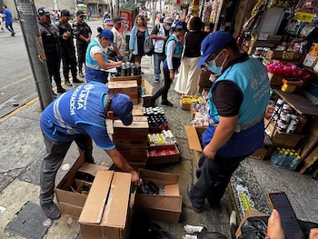 Funcionarios con chalecos azules revisan cajas de cartón con alimentos en una acera junto a transeúntes y agentes policiales
