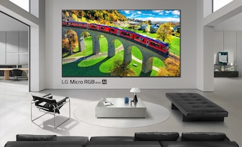 Televisor de gran tamaño en una pared blanca de un salón moderno con un sillón, una mesa redonda, un banco negro y un sofá