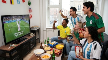 Un grupo de seis jóvenes sonríe y levanta latas de cerveza mientras mira un partido de la Copa Mundial en un televisor, con snacks en la mesa y banderas colgando.