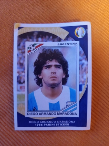 Estampilla cuadrada de Diego Armando Maradona con camiseta de Argentina. Incluye texto 'ARGENTINA' y '1986 PANINI STICKER', con iconos de fútbol y copa