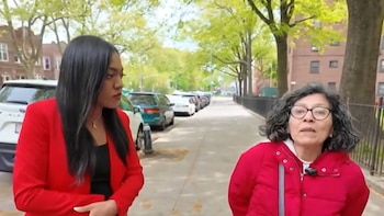 Dos mujeres conversando en una acera. La mujer de la izquierda lleva un blazer rojo y la de la derecha una chaqueta roja y gafas, con vehículos estacionados detrás