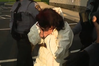 Un joven con traje de protección blanco se cubre el rostro con las manos mientras camina junto a oficiales de policía