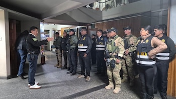 Un grupo de efectivos policiales vestidos con chalecos tácticos y de investigación escucha a un superior en el pasillo de un edificio