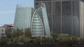 Vista exterior de un moderno edificio curvo con fachada de cristal junto a un rascacielos rectangular más alto, rodeados de árboles