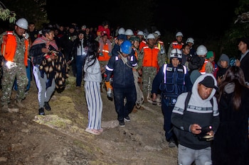 Varias personas, incluyendo civiles, personal militar y rescatistas con chalecos reflectantes y cascos, caminan por un camino de tierra oscuro usando linternas