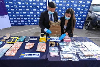 Dos agentes de la PDI con mascarillas y guantes inspeccionan una mesa llena de fajos de dinero chileno, joyas de oro, chaleco antibalas y teléfonos móviles