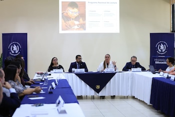 Varias personas en una mesa de reuniones con manteles. Una pantalla de proyección muestra un niño leyendo. Hay banderas de Guatemala y micrófonos sobre la mesa
