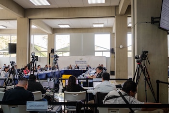 Integrantes de una comisión sentados en mesas en forma de U mientras camarógrafos y periodistas con computadoras cubren el evento