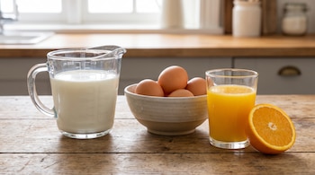 Una jarra de cristal con leche, un cuenco de cerámica con huevos, un vaso de jugo de naranja y media naranja sobre una mesa de madera.