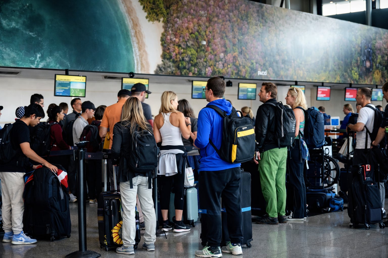 Un grupo de viajeros espera con su equipaje en una concurrida terminal de aeropuerto, con pantallas informativas y un gran mural paisajístico al fondo. (Imagen Ilustrativa Infobae)