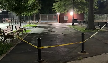 Un parque en Nueva York por la noche con cinta de seguridad amarilla bloqueando un camino, árboles, bancos de madera, farolas y un edificio