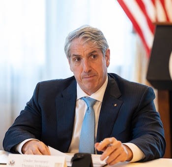 Thomas G. DiNanno, un hombre de cabello gris con traje oscuro y corbata azul, mira al frente sentado en una mesa con papeles, un bolígrafo y una bandera de EE. UU. al fondo