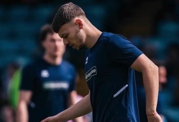 Primer plano de Jonathan Gjoshe, un jugador con camiseta azul oscuro con el logo Enterprise, mirando hacia abajo. Lleva una pulsera roja en la muñeca izquierda