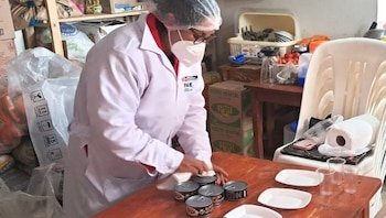 Una mujer con bata blanca, mascarilla y cofia manipula latas de conserva sobre una mesa de madera junto a platos y vasos de plástico