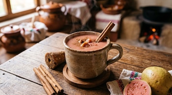 Una taza humeante de atole con una rama de canela, junto a canela en rama, un cono de piloncillo y guayabas sobre una mesa de madera.
