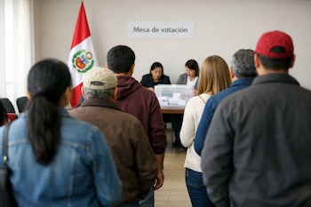 Una fila de personas de espaldas espera para votar en un consulado. Se observa una bandera peruana, una mesa de votación con urna y dos funcionarios electorales.