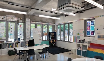Un aula espaciosa con paredes blancas y techo de hormigón, grandes ventanas que muestran vegetación, mesas blancas y sillas negras, y materiales educativos en las paredes
