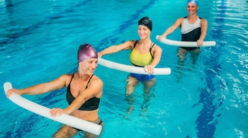 Tres mujeres caucásicas con gorros y trajes de baño en una piscina azul, sosteniendo flotadores cilíndricos de espuma blanca.