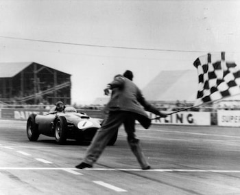 Coche de carrera número 7 con piloto en pista. Un oficial ondea bandera a cuadros. Carteles publicitarios y graderías al fondo